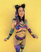 Vibrant and joyful raver in colorful psychedelic attire with hood and fluff ears poses against a sunny yellow wall.