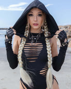 Woman wearing a matte black assassin hood and a stylish cut-out swimsuit at the beach, showcasing a trendy festival look.