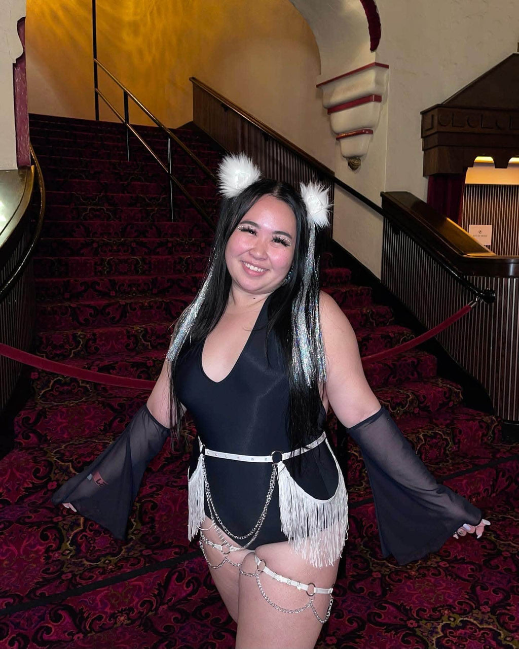 Young woman in a black bodysuit with fringe and accessories, posing on a grand staircase, showcasing rave outfit style.