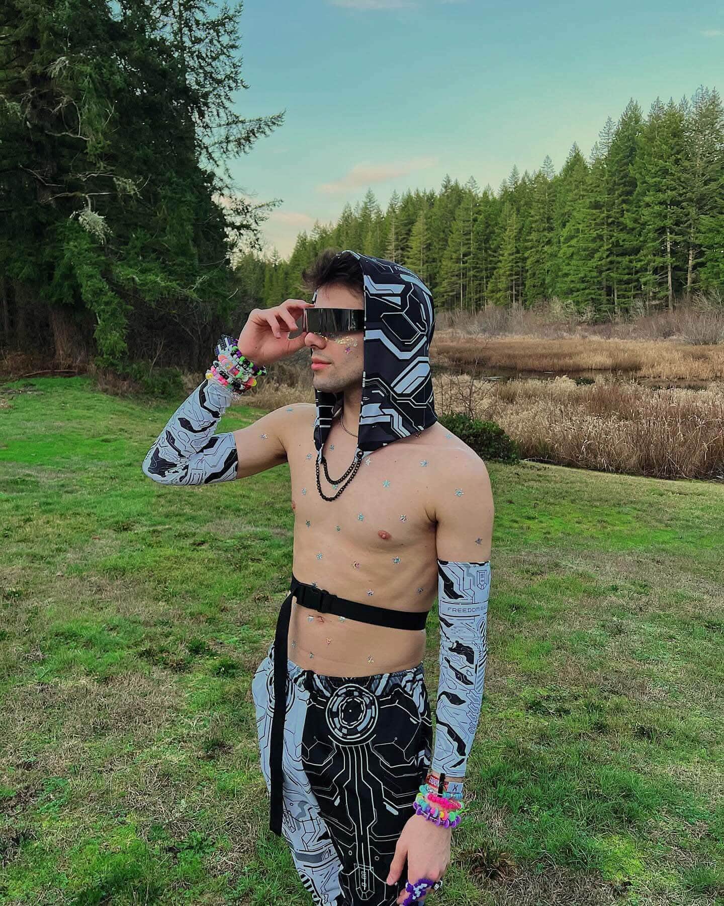 Young man in a patterned hoodie and arm sleeves poses outdoors, showcasing a stylish rave outfit among trees.