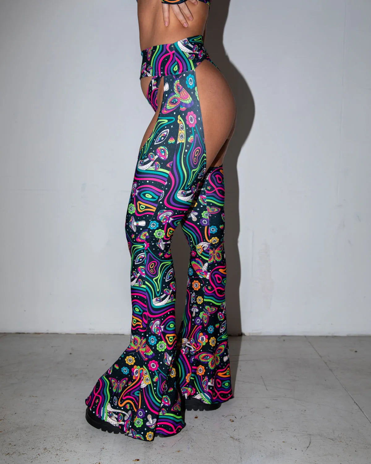 Model in Awaken Bell Chaps, side profile, showcasing the vibrant psychedelic print with mushrooms, eyes, and butterflies, and the open hip design. Freedom Rave Wear.