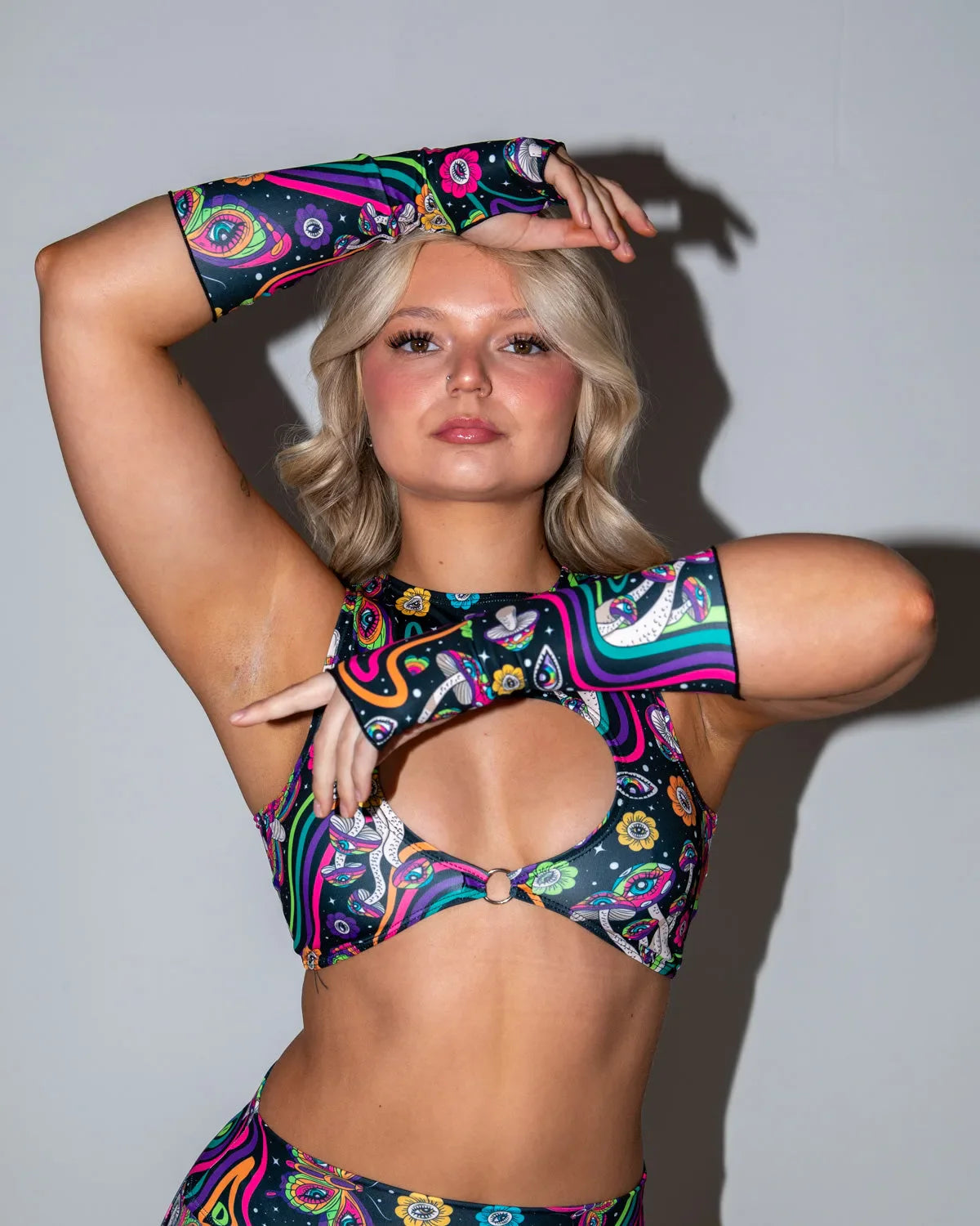 Awaken Gloves and matching top, featuring a vibrant psychedelic print with eyes, mushrooms, and wavy lines. Model poses with arms raised, waist-up shot. Freedom Rave Wear.