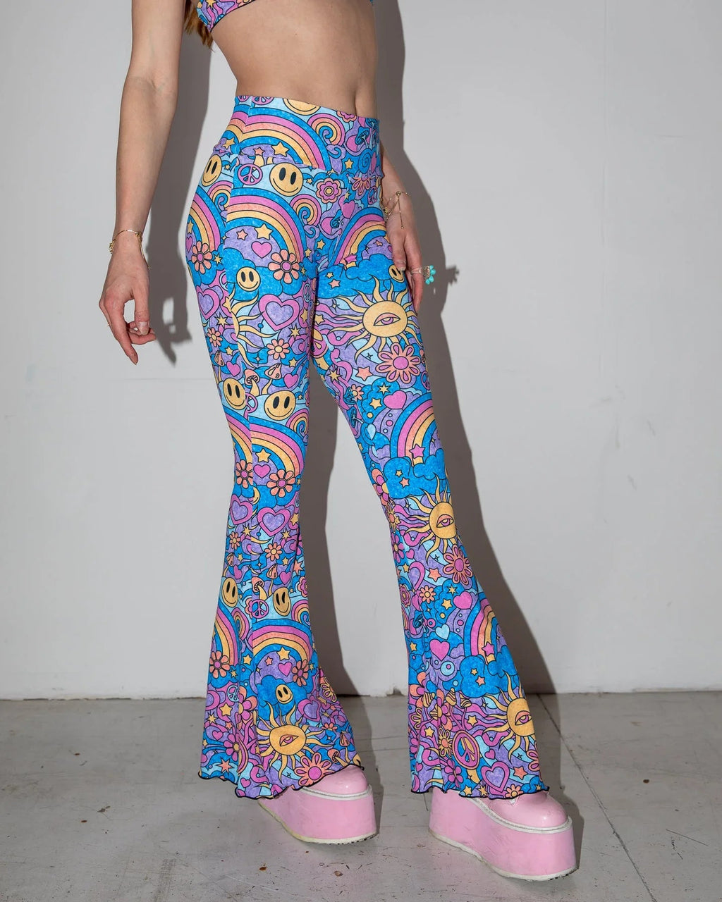 Groovy Bell Bottoms in a vibrant blue and purple psychedelic pattern, featuring smiley faces, peace signs, and rainbows, worn by a model from the front, perfect for rave festivals.