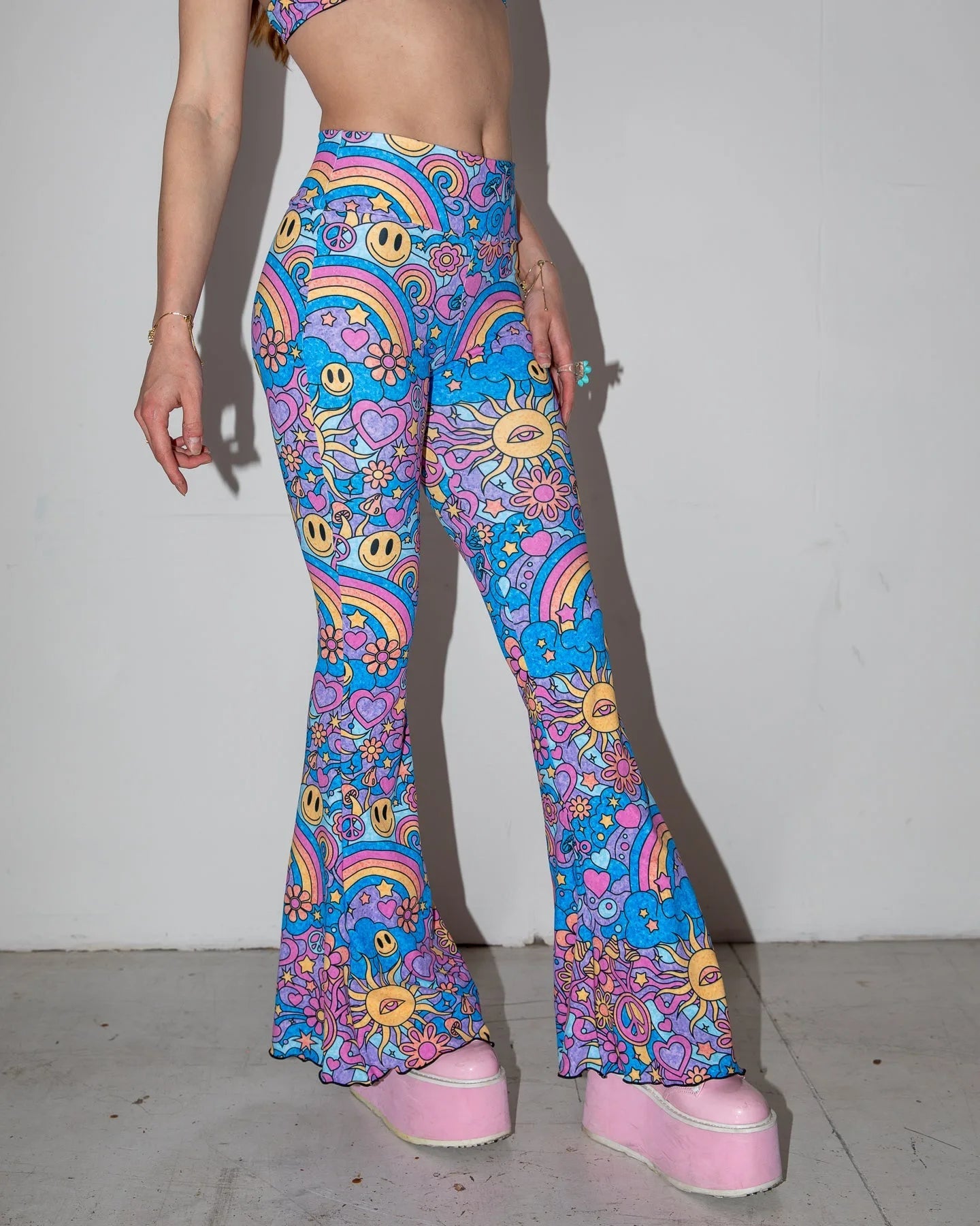 Groovy Bell Bottoms in a vibrant blue and purple psychedelic pattern, featuring smiley faces, peace signs, and rainbows, worn by a model from the front, perfect for rave festivals.