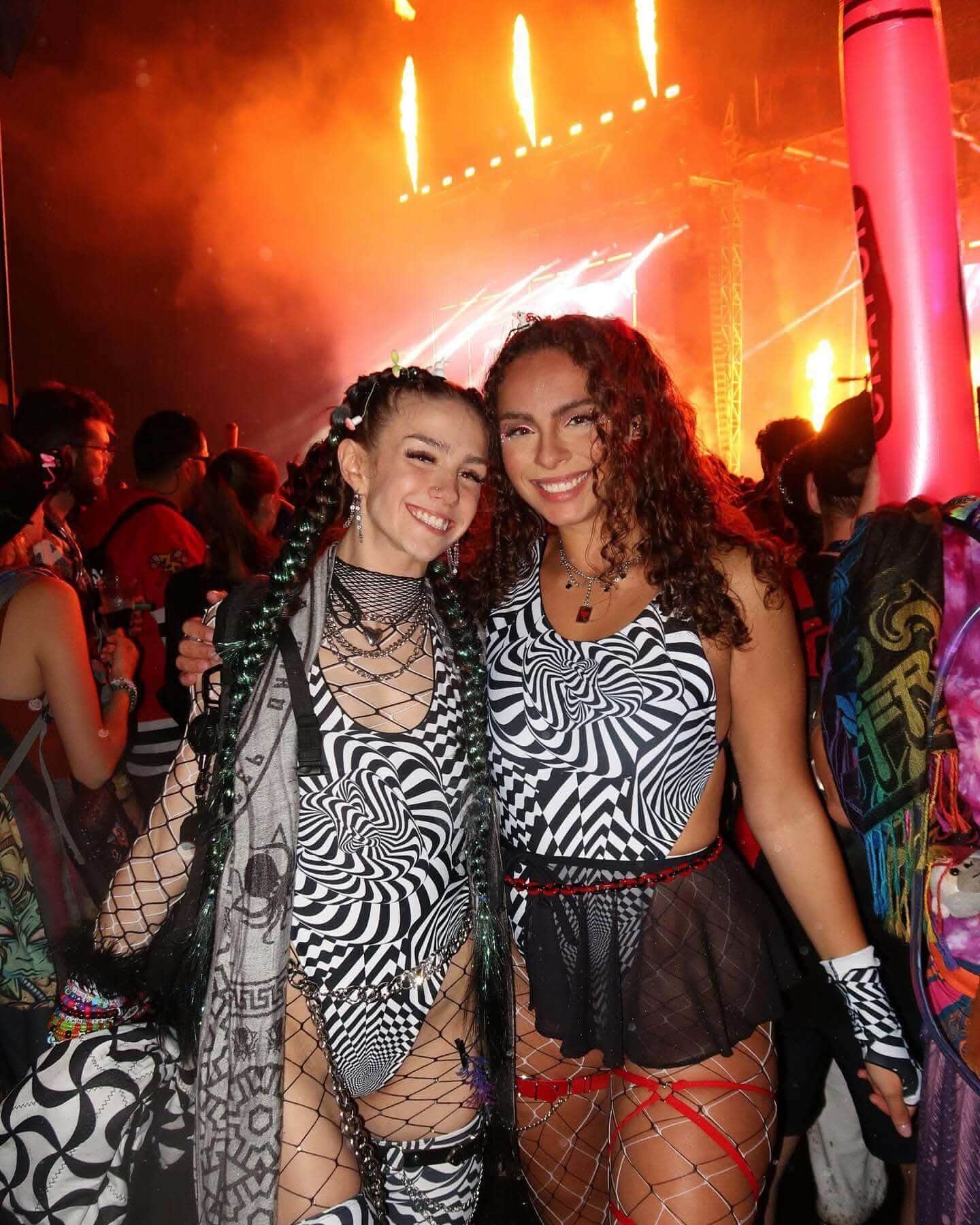 Two festival-goers wearing Distortion print rave bodysuits at EDM event with dramatic stage lighting