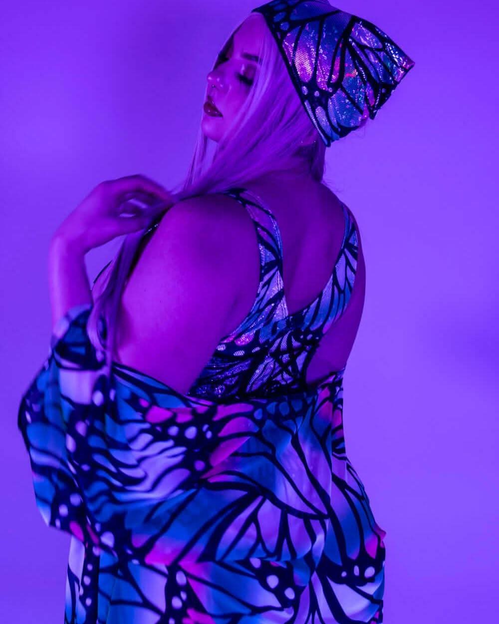Model wearing Flutter butterfly pattern rave bodysuit and matching bandana headwrap in purple neon festival lighting