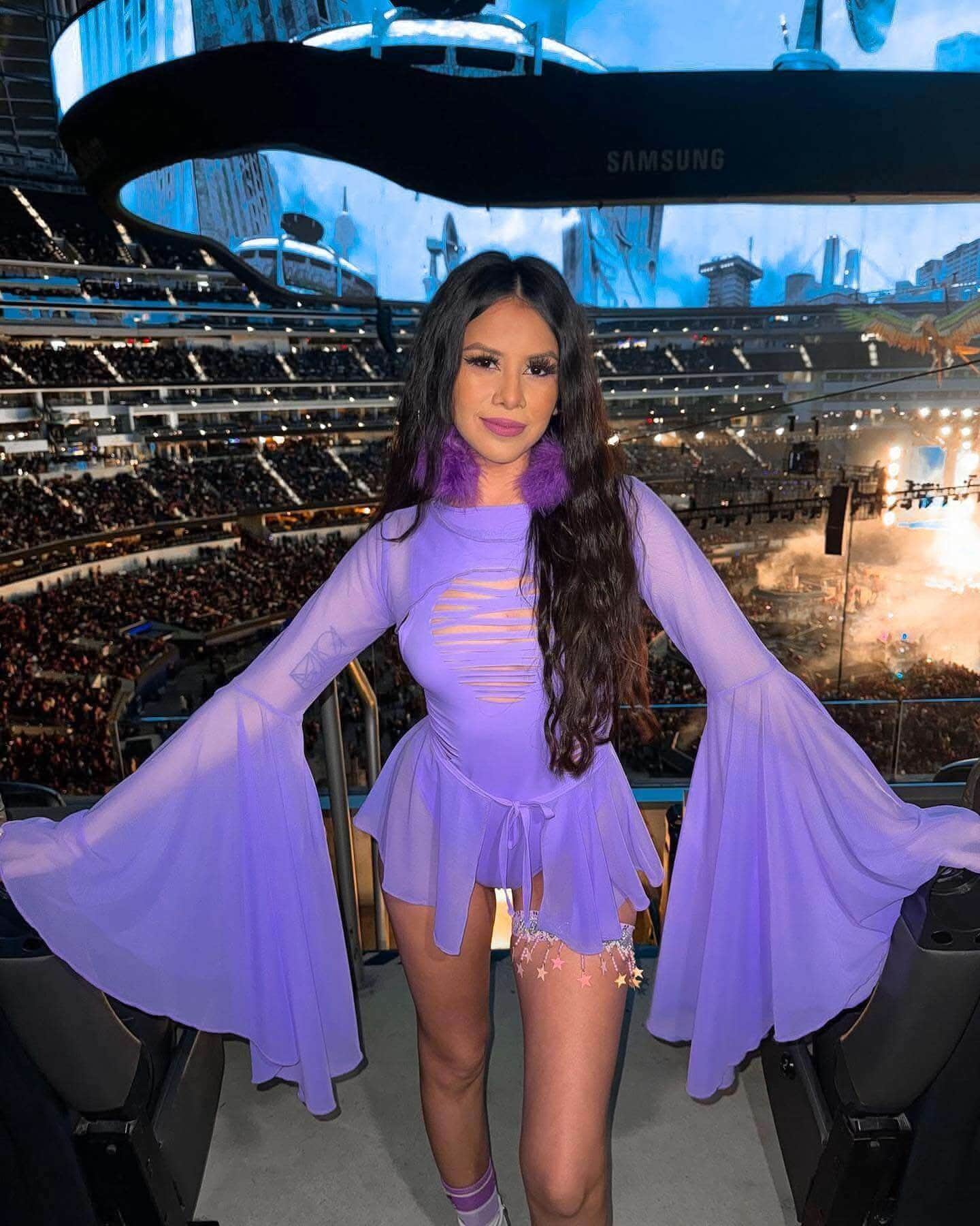 Model wearing lavender rave bodysuit with bell sleeves posing at festival venue with stadium crowd background