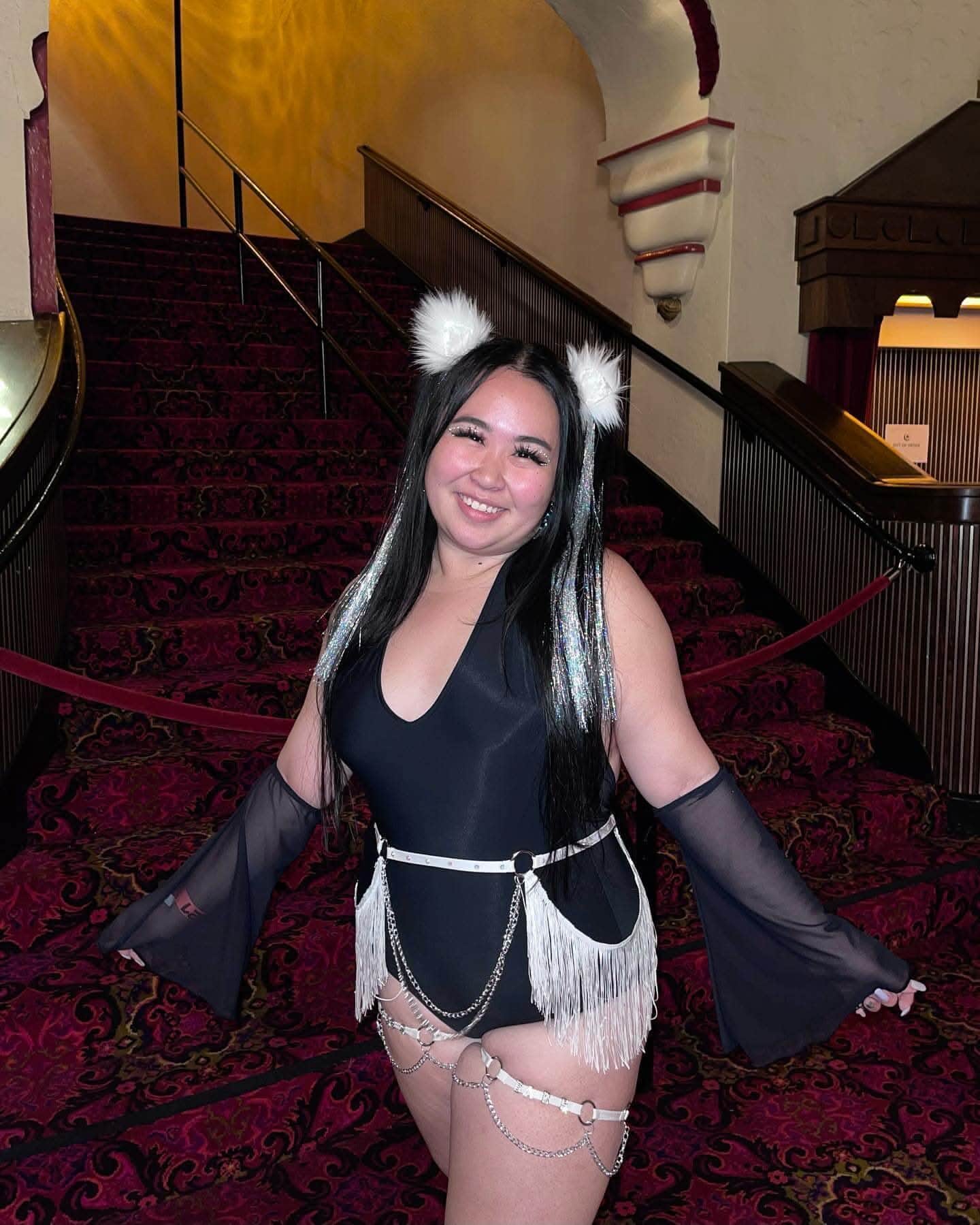 Model wearing matte black rave bodysuit with white fringe belt on festival staircase pose