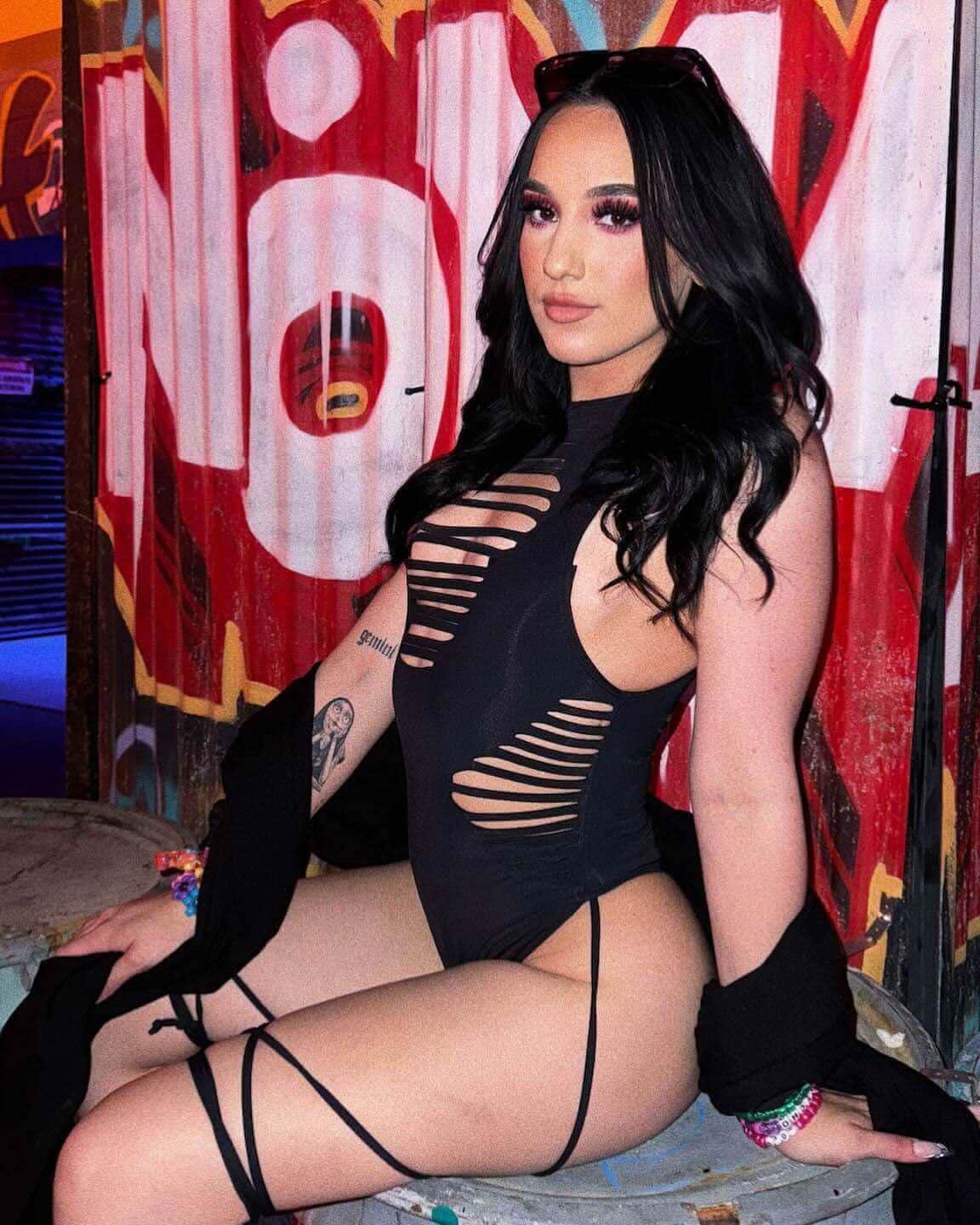 Model in matte black rave bodysuit with sideboob slits and leg straps against colorful graffiti wall