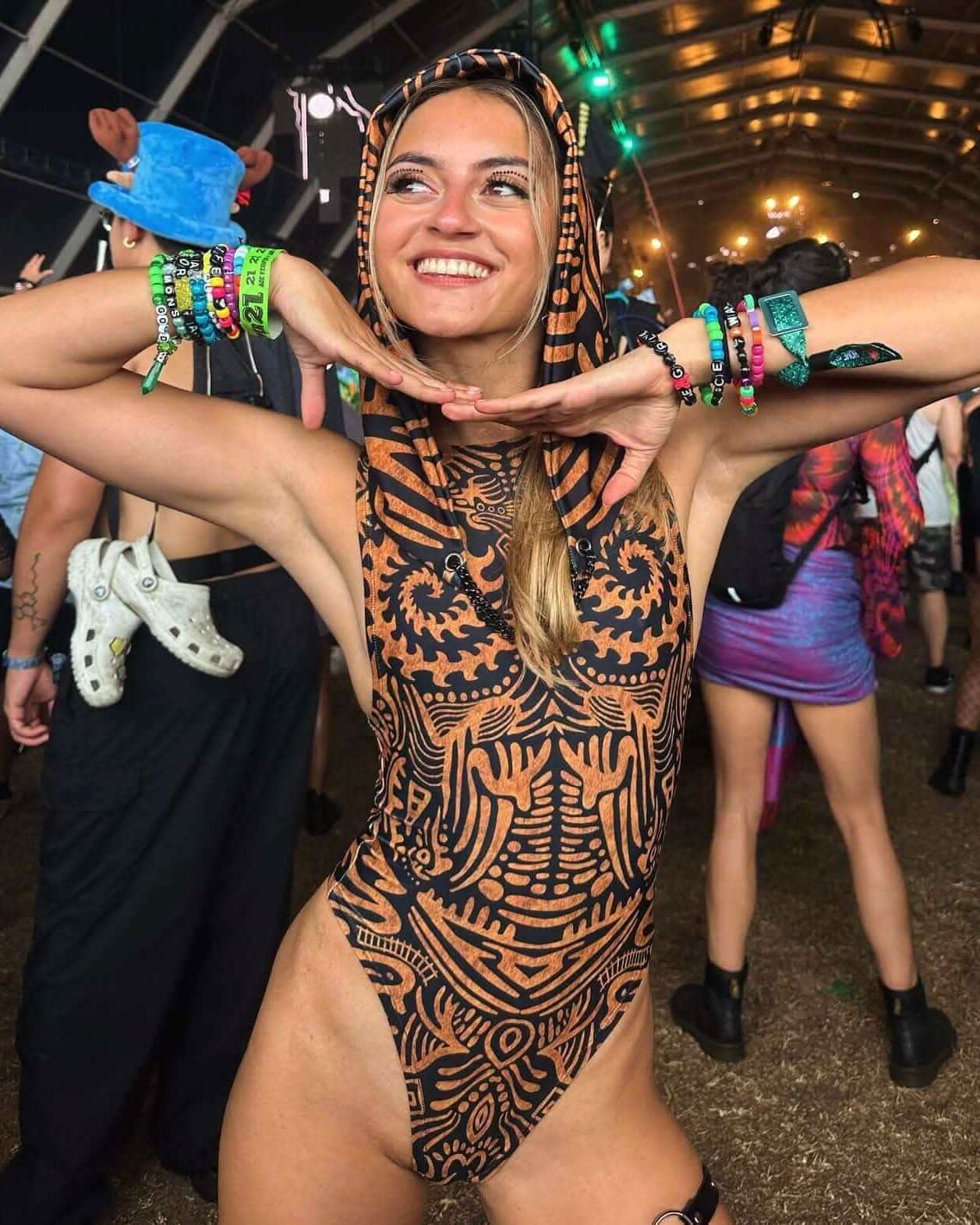 Woman wearing orange black Primal rave bodysuit with hood posing at festival event with stage lights