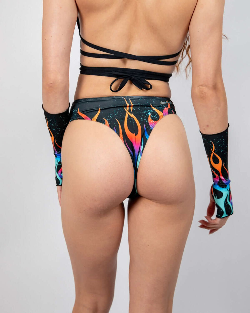 Model from behind wearing the Blaze High Waisted Thong, showcasing its vibrant neon flame pattern and teal speckles, perfect for a rave festival.