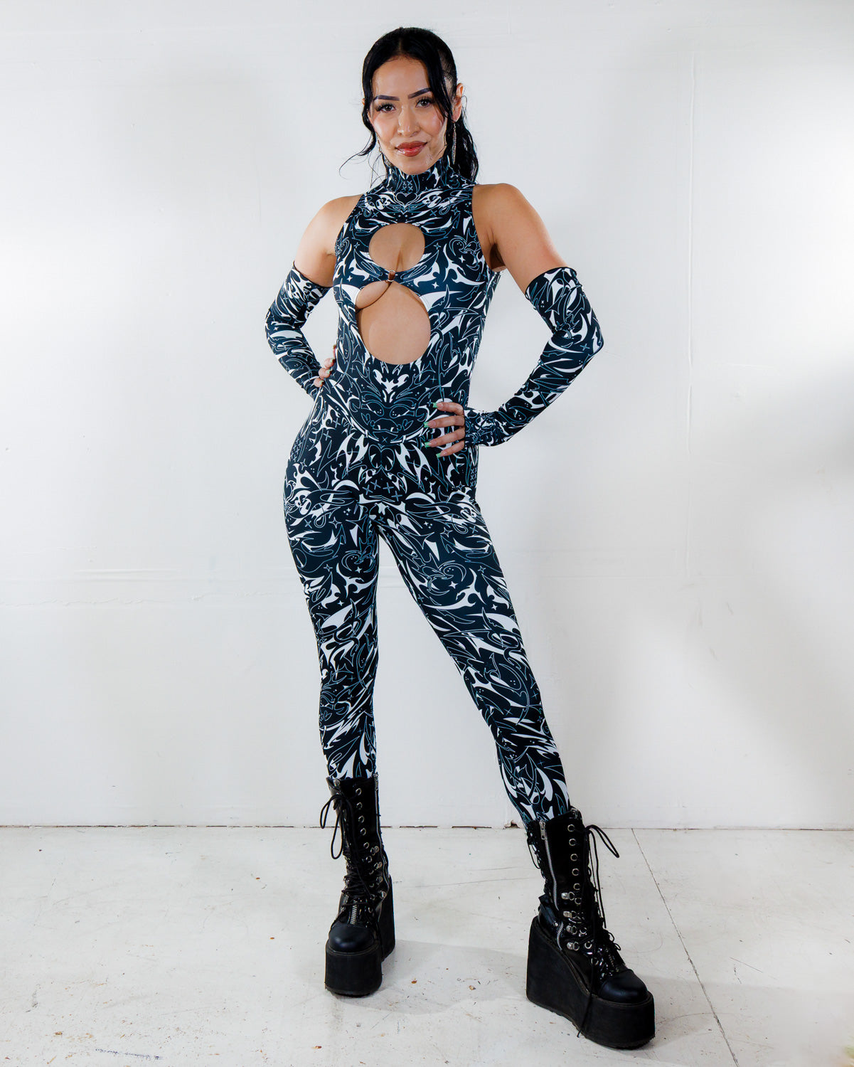 Model in BLVDE Cold Shoulder Jumpsuit, a black and white patterned rave bodysuit with chest and midriff cutouts, posing front-facing.