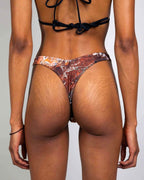 Back view of model wearing Fossil Risk rave pants with brown prehistoric pattern and black strappy top