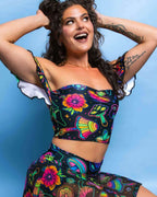 Smiling model in colorful Shroomie mushroom print rave top with off-shoulder ruffles and sweetheart neckline against blue background