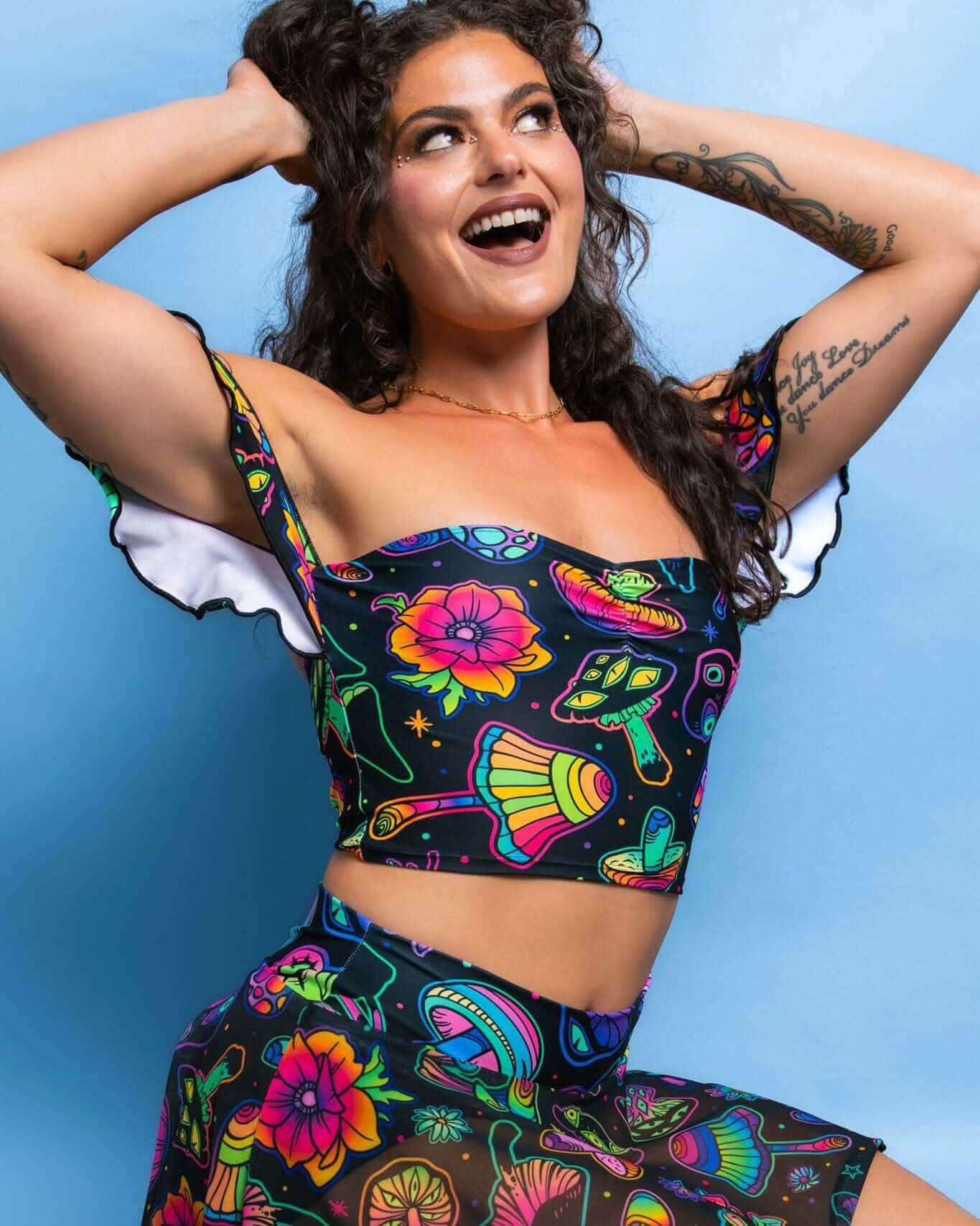 Smiling model in colorful Shroomie mushroom print rave top with off-shoulder ruffles and sweetheart neckline against blue background