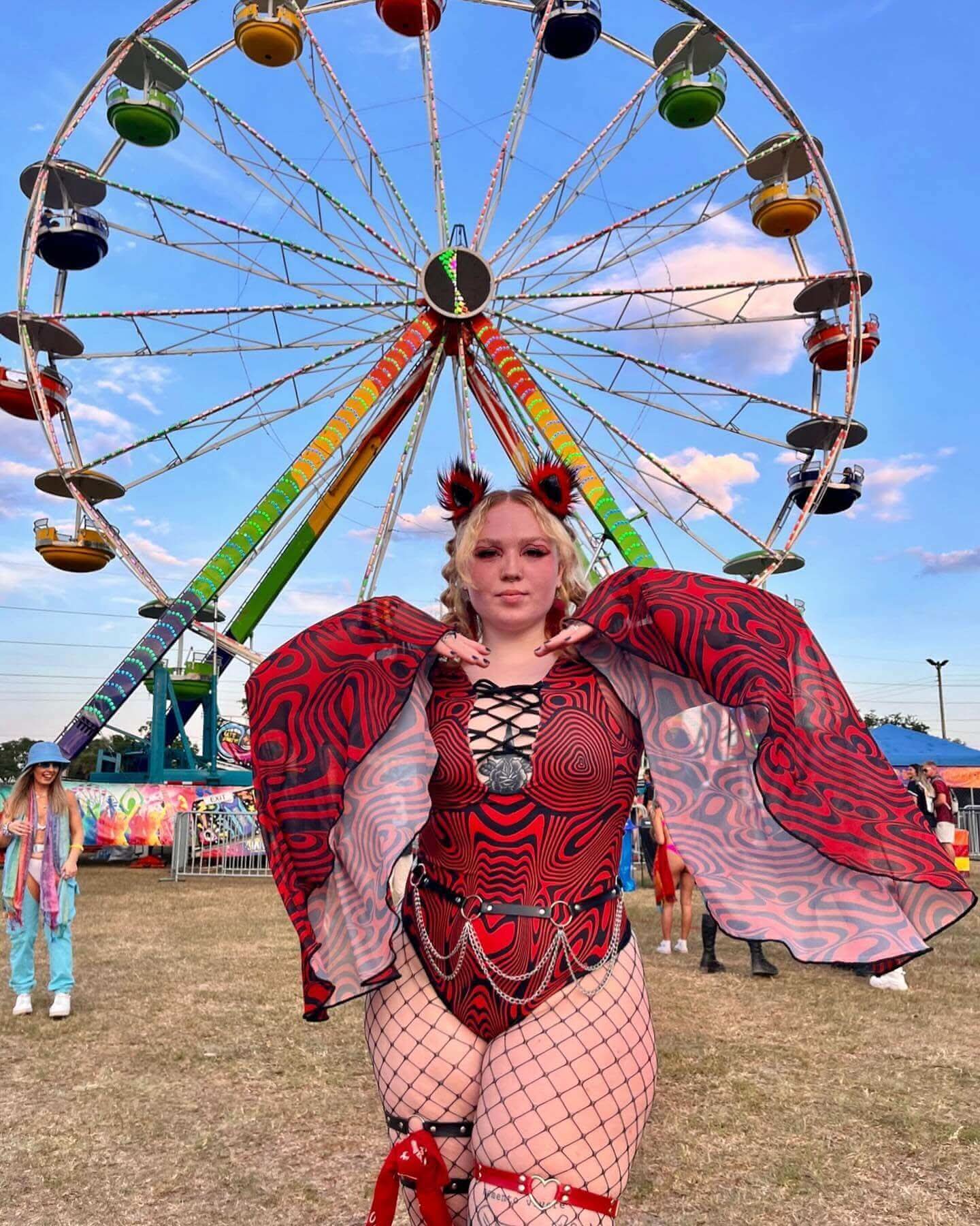 Woman in a vibrant Red Void Goddess Bodysuit poses in front of a colorful ferris wheel, showcasing rave outfits.