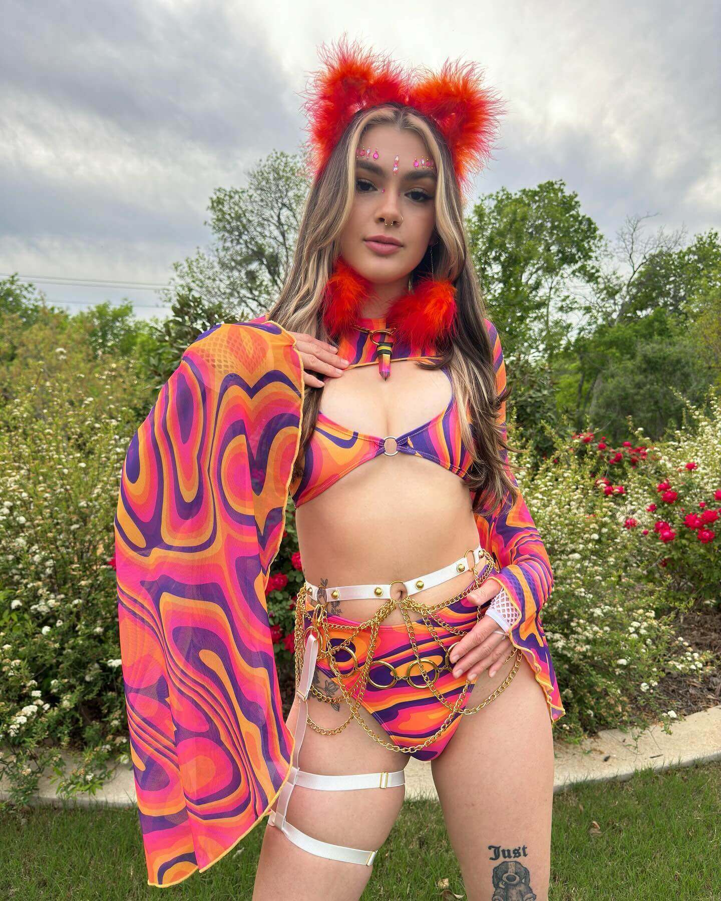 Model wearing a vibrant orange and purple psychedelic rave outfit with bold accessories in a garden setting.