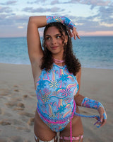 Person on the beach in a psychedelic blue and pink floral one-piece, matching sleeves and choker, with a pink chain belt.