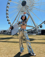 Raver in futuristic metallic outfit with fan and visor glasses stands poised before a giant Ferris wheel under a clear blue sky.