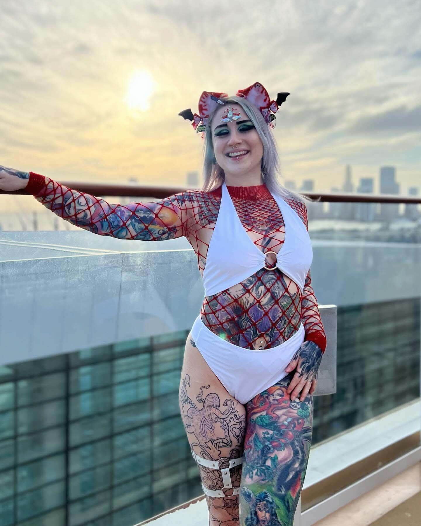 Woman in a colorful rave outfit poses with a city skyline and sunset in the background, showcasing unique body art and style.
