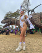 Stunning model in white eco-luxe metamorphosis arm sleeves, posing in front of a dinosaur statue at a lively outdoor event.
