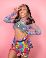 Smiling woman in vibrant patterned outfit posing playfully against a pink background, perfect for rave outfits.