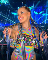 Woman wearing colorful rave outfit with rainbow arm sleeves, smiling at a vibrant event with bright lighting.