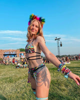 Woman in a trippy rainbow bralette and accessories, showcasing rave outfits at a music festival in a grassy field.