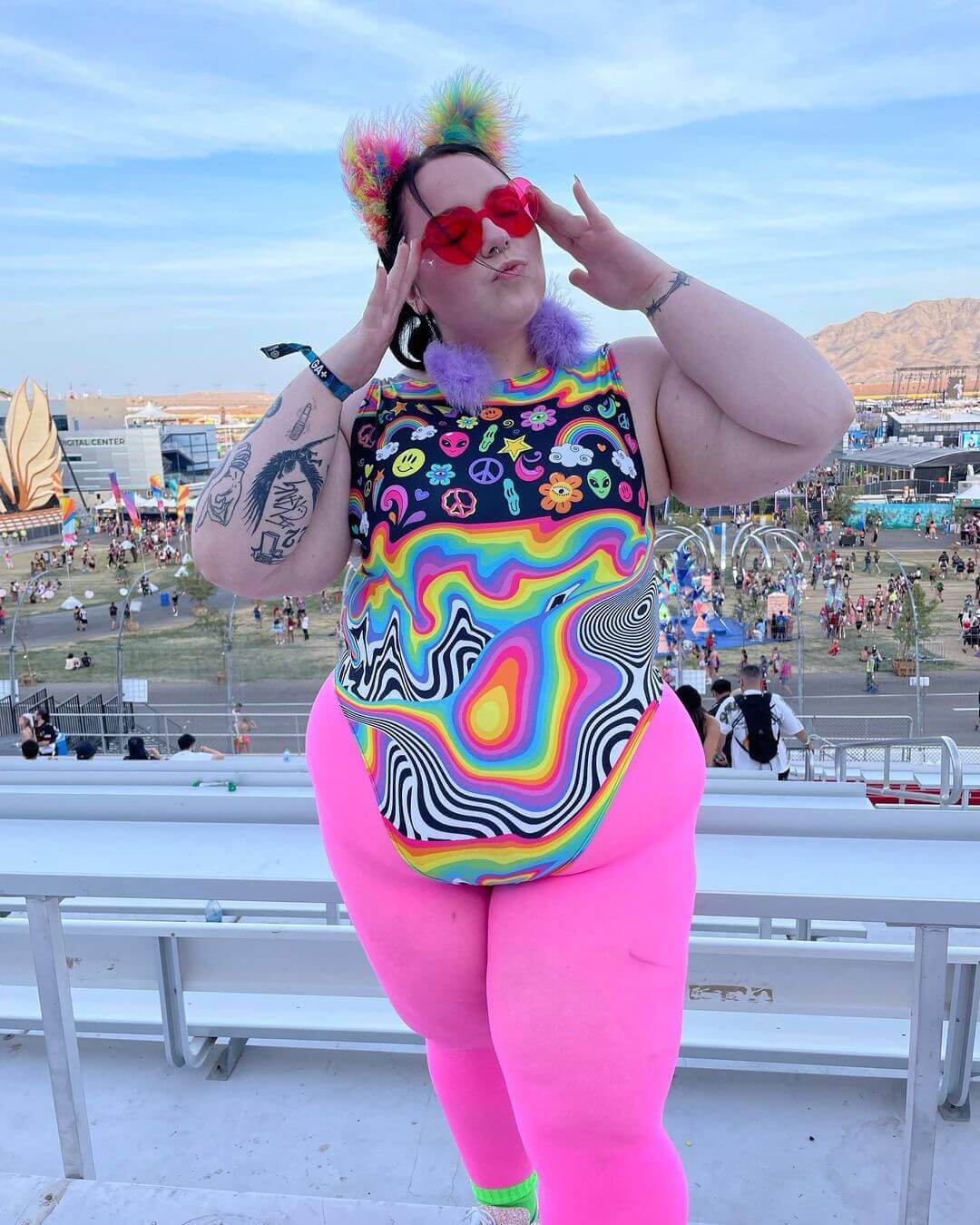 Trendy plus-size model in a colorful rainbow sideboob bodysuit at a vibrant EDM festival, showcasing bold rave outfits.