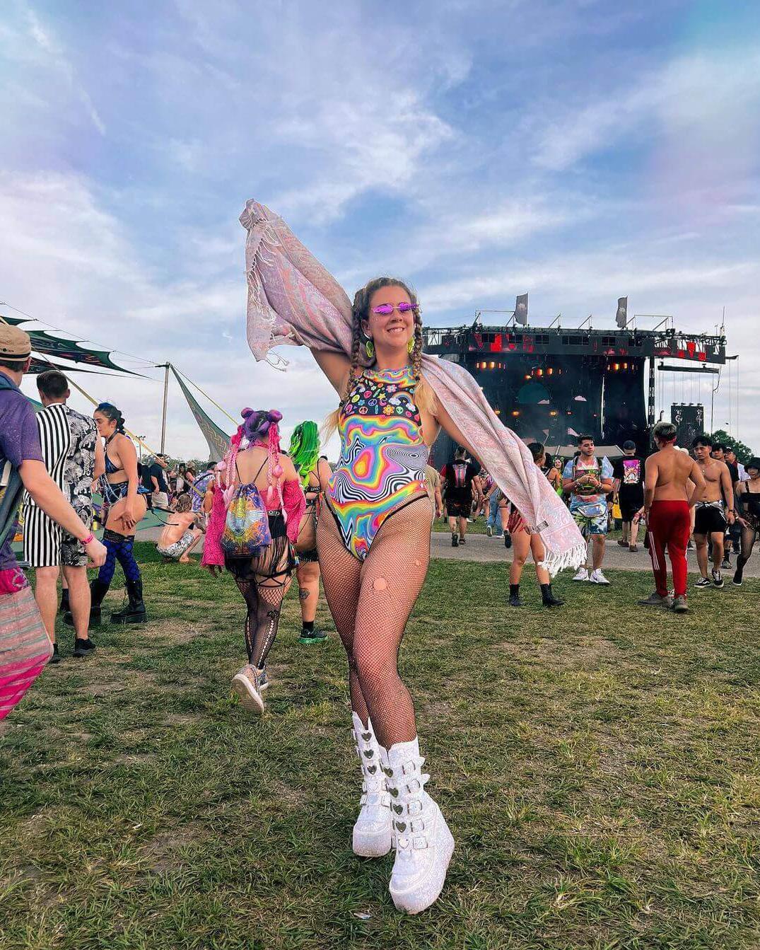 Vibrant woman in psychedelic rainbow bodysuit at EDM festival, showcasing trendy rave outfits and summer festival fun.