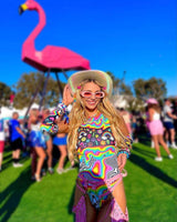 Woman wearing a psychedelic rainbow bodysuit at a festival, surrounded by colorful outfits and a pink flamingo backdrop.
