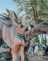 Woman in a rave outfit poses with a dinosaur statue at a festival, surrounded by colorful crowd and greenery.