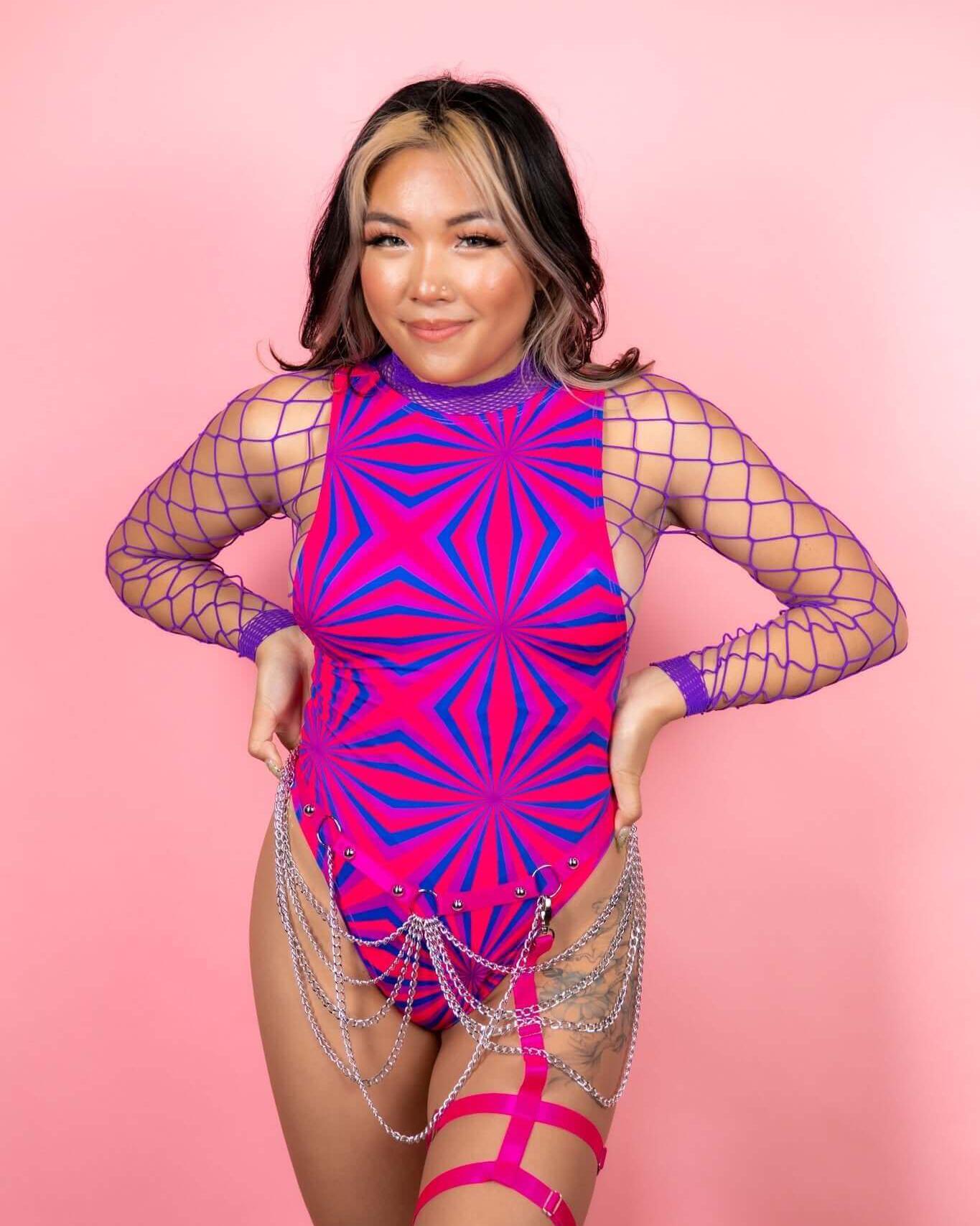 Woman in vibrant pink bodysuit with geometric patterns and chain details, styled for rave outfits against a pink background.