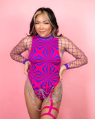 Woman in vibrant pink bodysuit with geometric patterns and chain details, styled for rave outfits against a pink background.