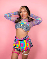 Smiling model in colorful rave outfit with pink leather garter, against a pink background, showcasing festival fashion.