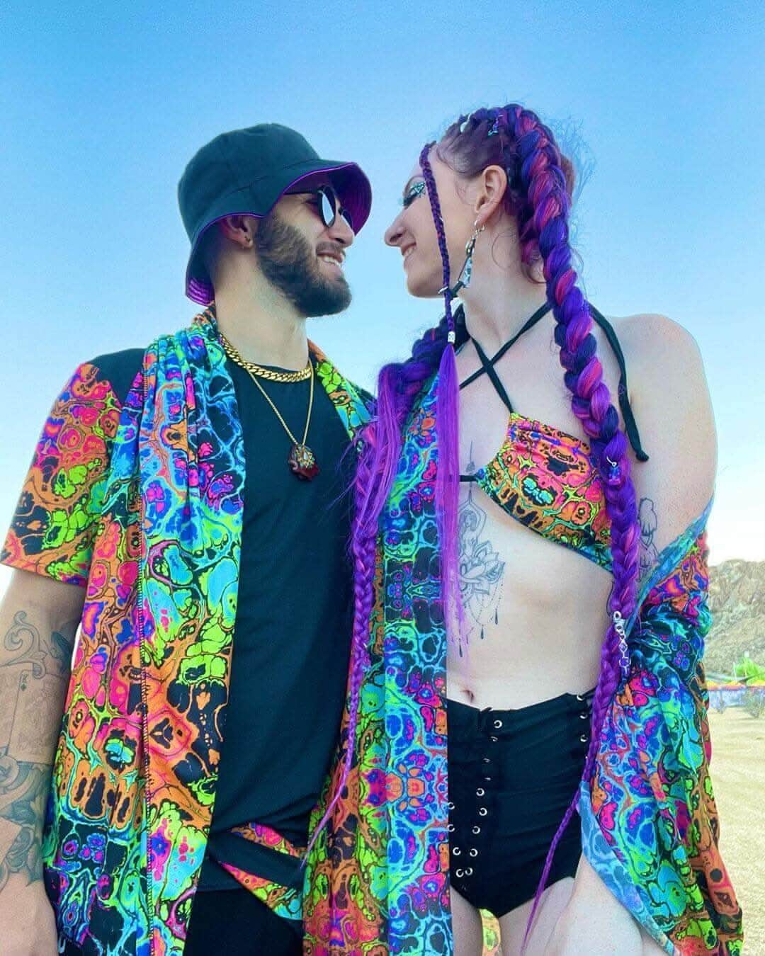 A couple wearing vibrant, psychedelic rave outfits, smiling at each other at a festival under a clear blue sky.