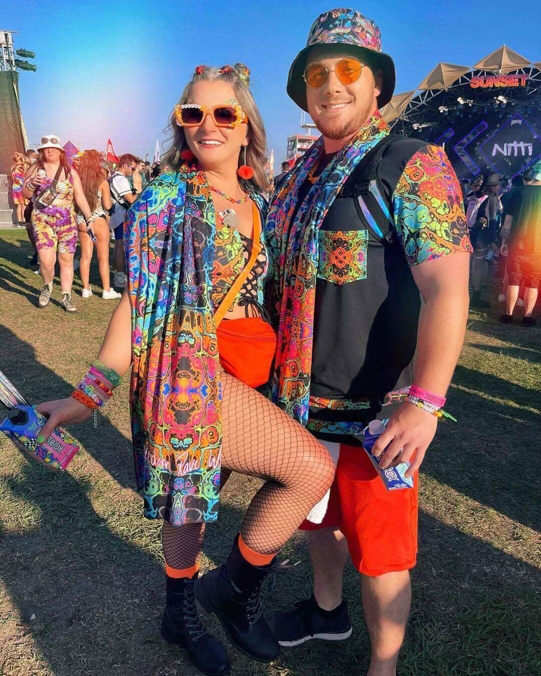 Colorful couple in vibrant rave outfits and accessories at a lively music festival with a sunny backdrop.