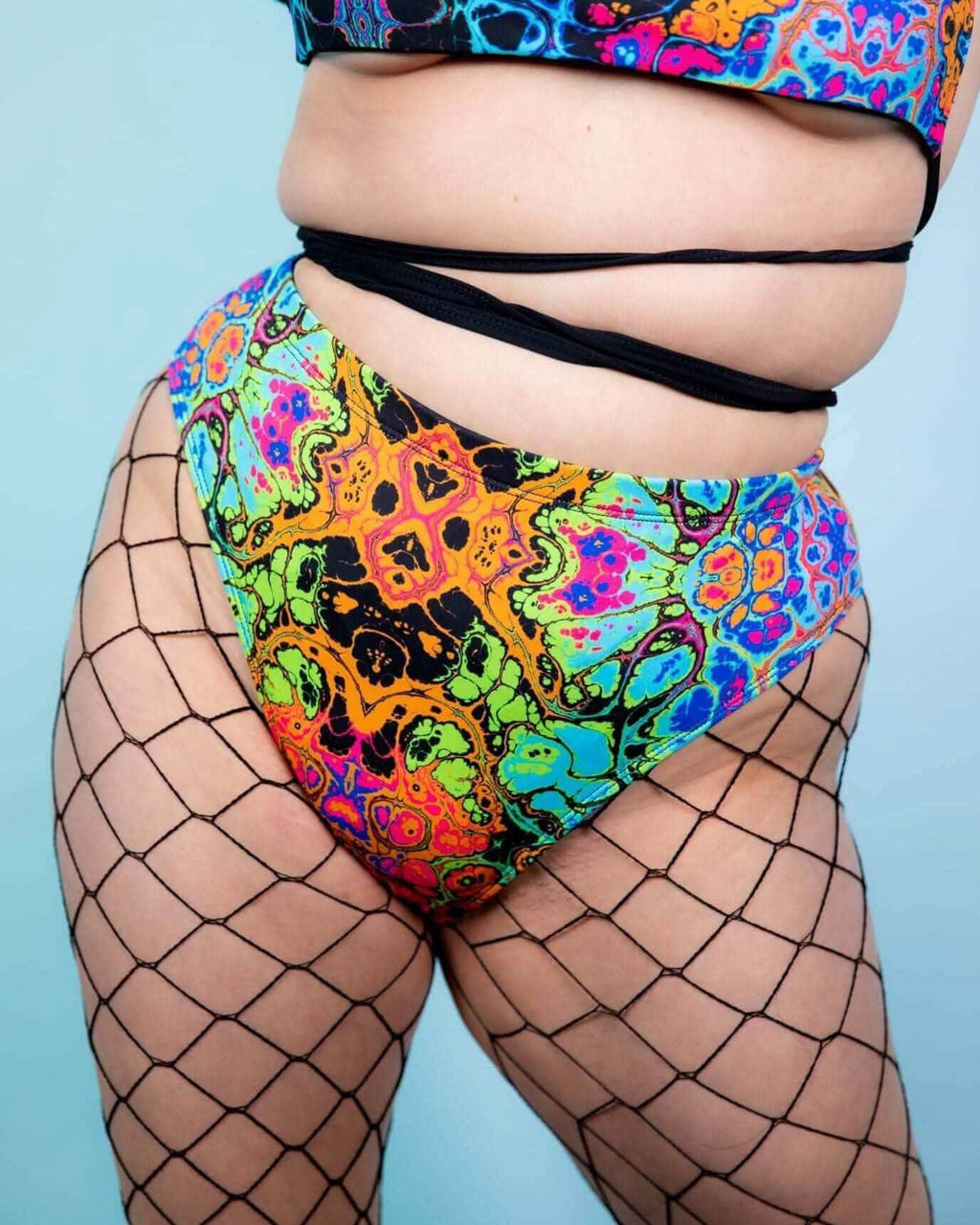 Model wearing colorful Lucidity High Waisted Brazilian Bottoms with fishnet tights against a light blue background.