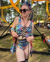 Rave girl in a colorful psychedelic rave two piece relaxes around green foliage , sunglasses on, exuding festival vibes