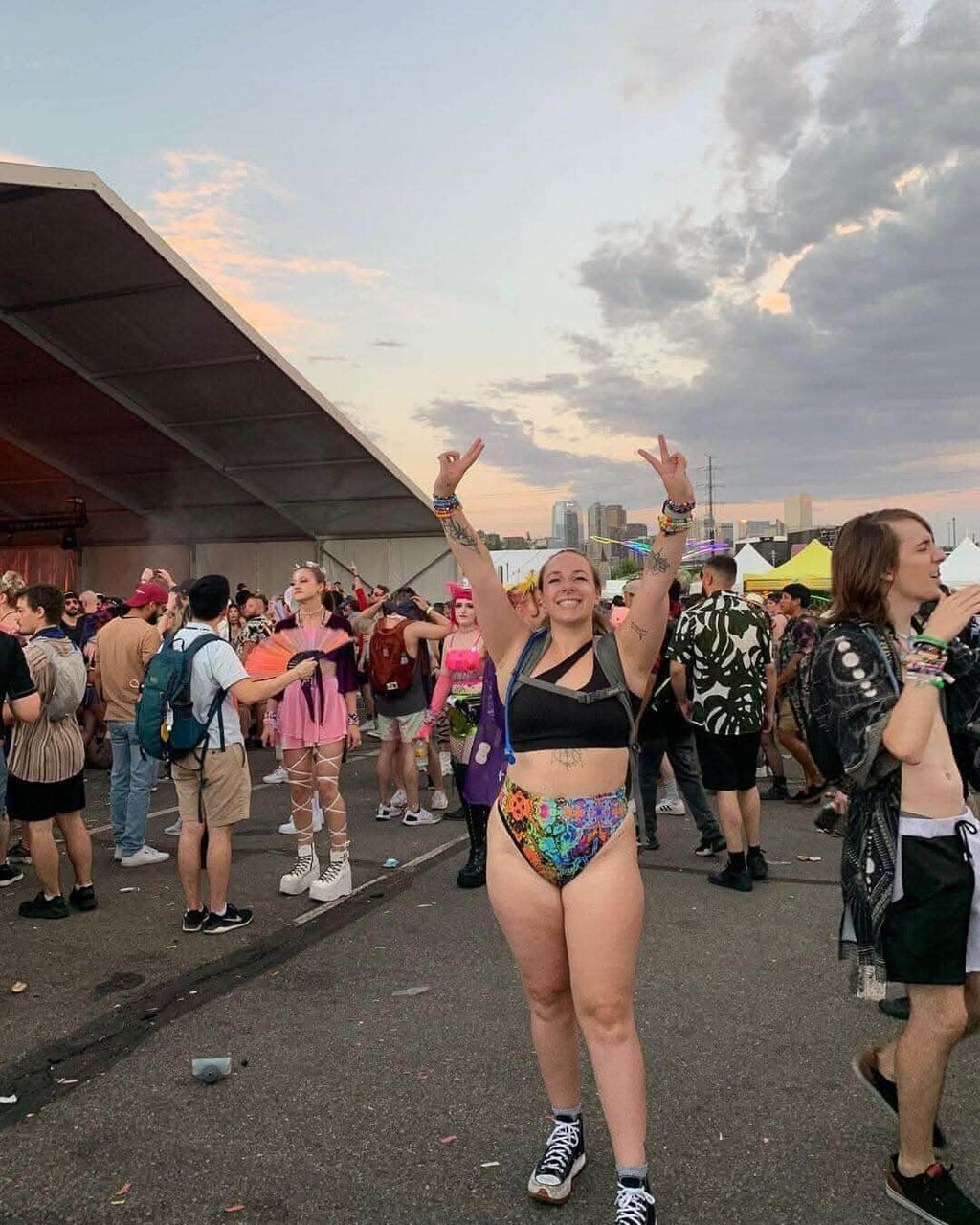 Woman in psychedelic high-waisted thong at a festival, surrounded by a lively crowd, celebrating with peace signs.