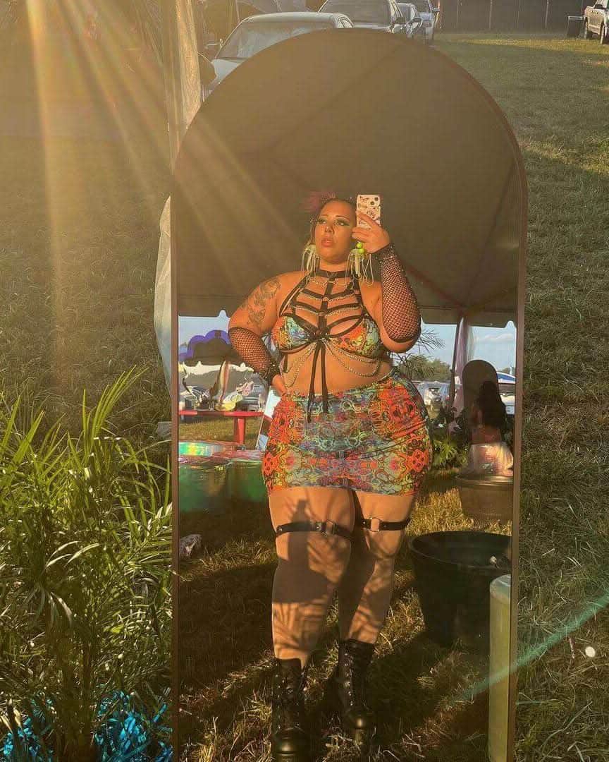 Woman in a colorful rave outfit with mesh details standing in front of a mirror at an outdoor festival.