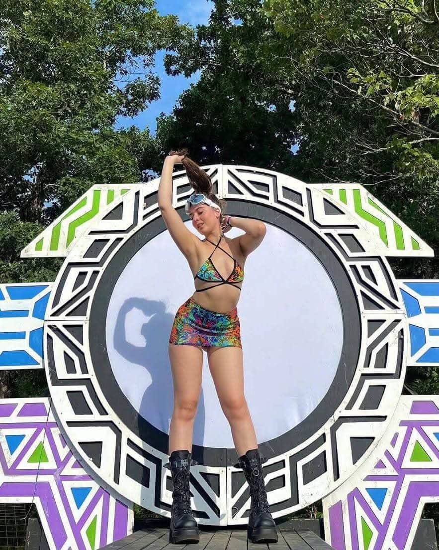 Woman modeling psychedelic rainbow rave outfit in front of colorful stage backdrop, showcasing vibrant festival style.