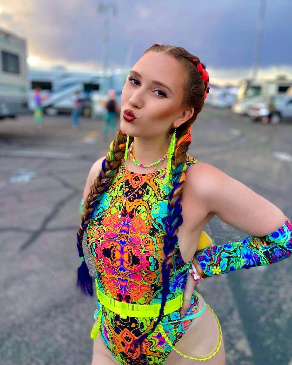 Colorful woman posing in a neon high-neck bodysuit, showcasing vibrant psychedelic print and bold accessories at a festival.