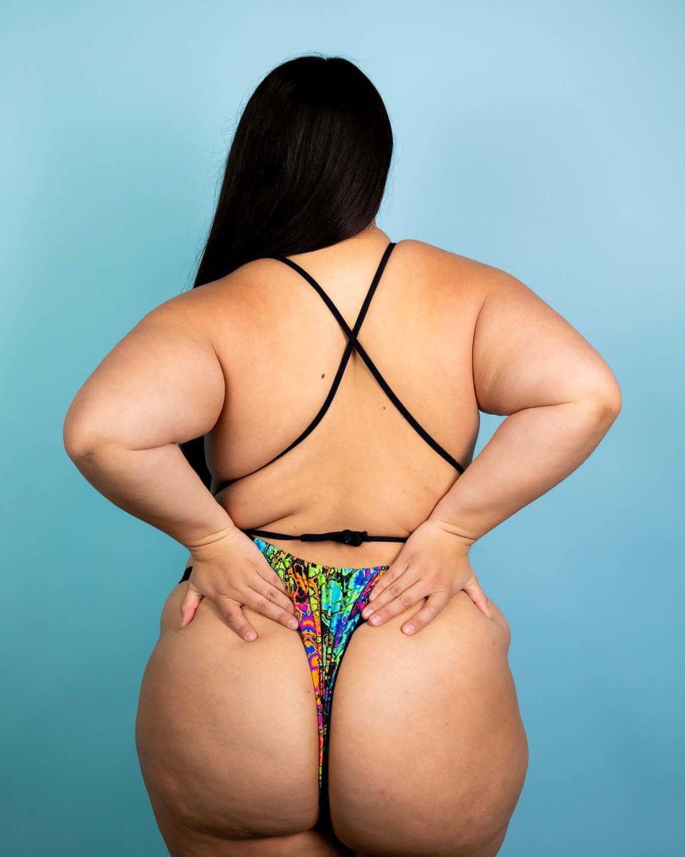 Back view of a model wearing the Lucidity Temptation Cheeky One Piece, showcasing its vibrant rainbow design and minimal coverage.