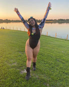Woman in a black one-piece swimsuit with colorful sleeves, joyfully celebrating by the water at sunset.
