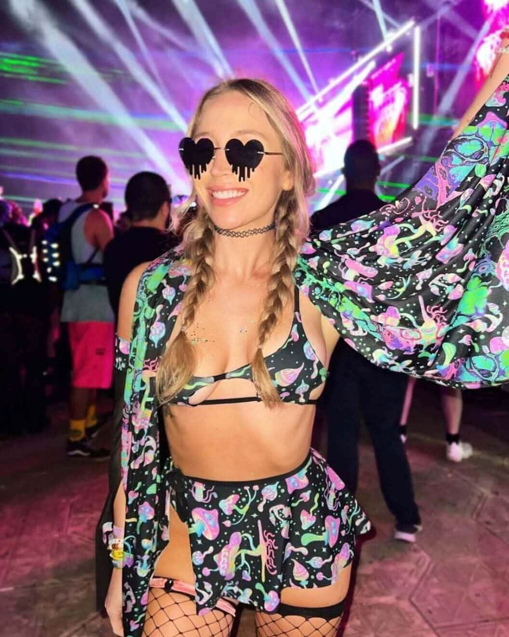 Woman wearing a colorful rave outfit with a mini skirt, sunglasses, and a wrap, enjoying a vibrant music festival atmosphere.