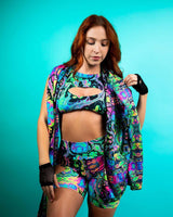 Vibrant woman modeling prismatic rave outfit with colorful top, shorts, and scarf against a teal background.