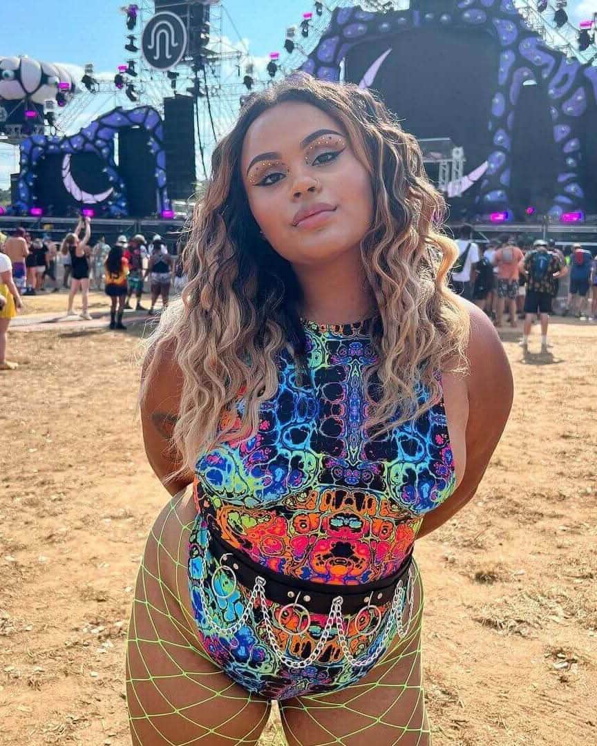 Woman wearing Lucidity psychedelic rainbow rave bodysuit at outdoor music festival with stage backdrop