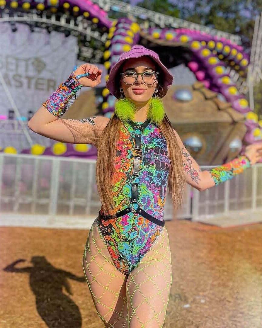 Woman wearing Lucidity psychedelic rainbow rave bodysuit with sideboob cutouts and green fur trim at outdoor festival