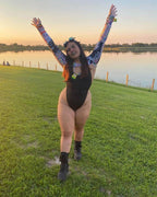Woman wearing matte black rave bodysuit with colorful arm sleeves celebrating at sunset festival field